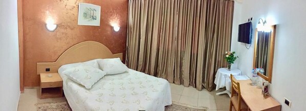 A bedroom with a bed, a bedside table, a desk, a chair, a mirror, a TV, and curtains. - Hotel Yasmine (Sfax)