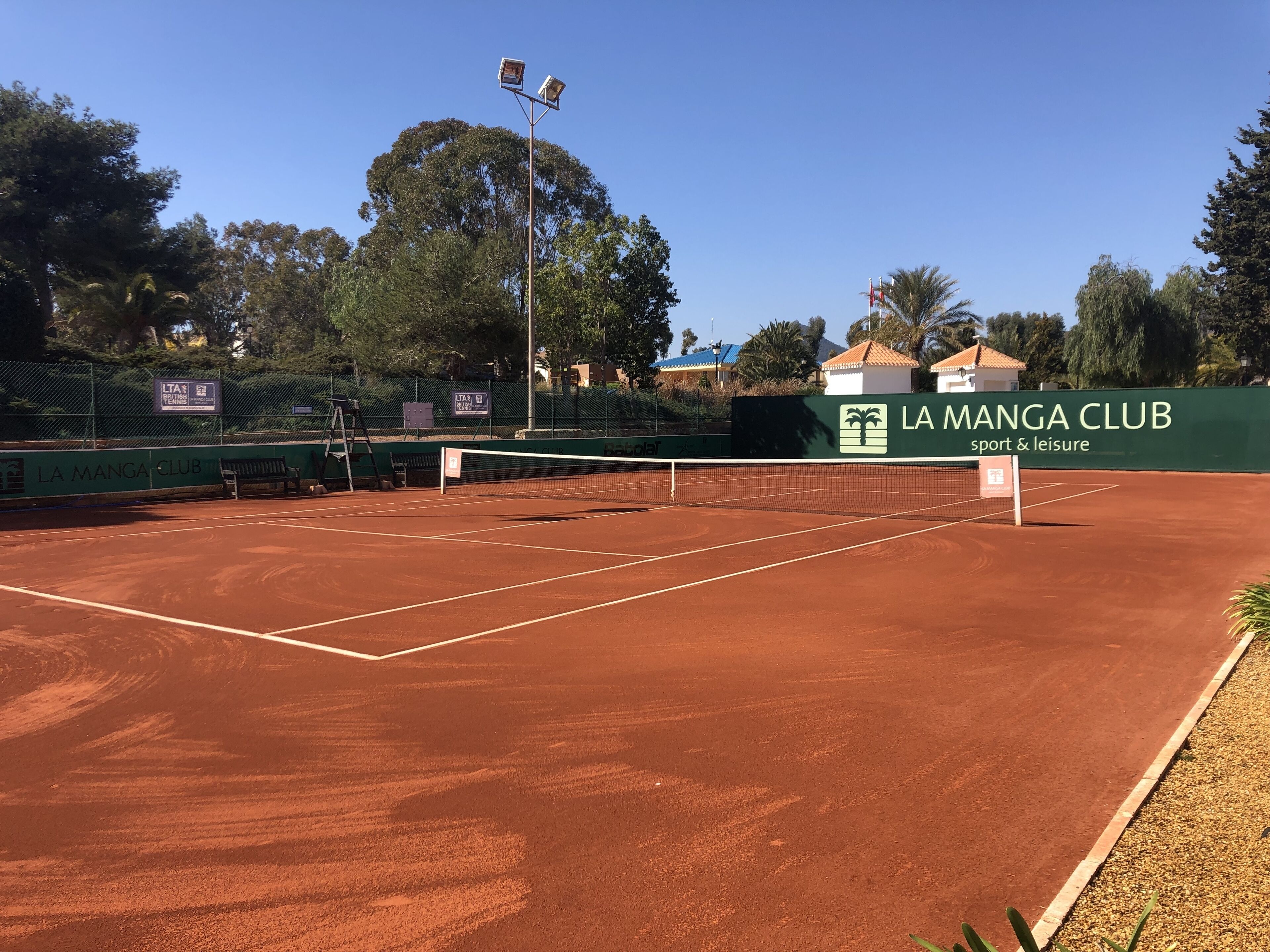 A tennis court with a net, surrounded by a fence, and a sign for La Manga Club.
