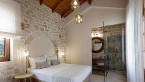 A bedroom with a stone wall, a wooden ceiling, a bed with pillows, a small stool, and a glass-enclosed shower area. - Superb villa Rogdia with heated pool, 2 minutes walking from local amenities (Rethymno)