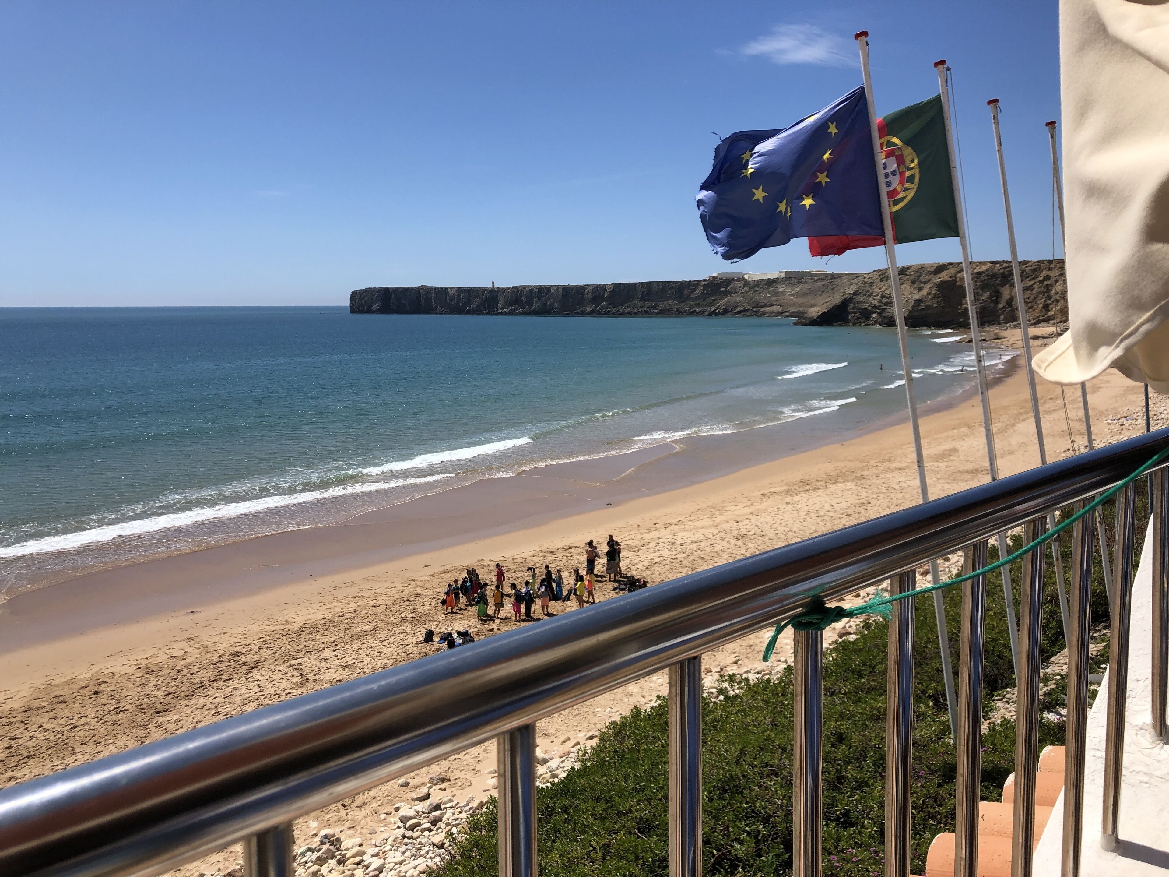 A beach with flags, a railing, and a rocky cliff in the background.