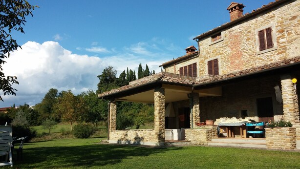 Une maison en pierre dotée d’un patio couvert, d’une cheminée en pierre et d’un jardin avec une pelouse et des arbres.