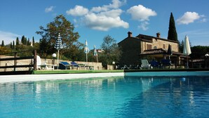Une piscine entourée d’un bâtiment en pierre et d’arbres, avec des chaises longues et des parasols à proximité.