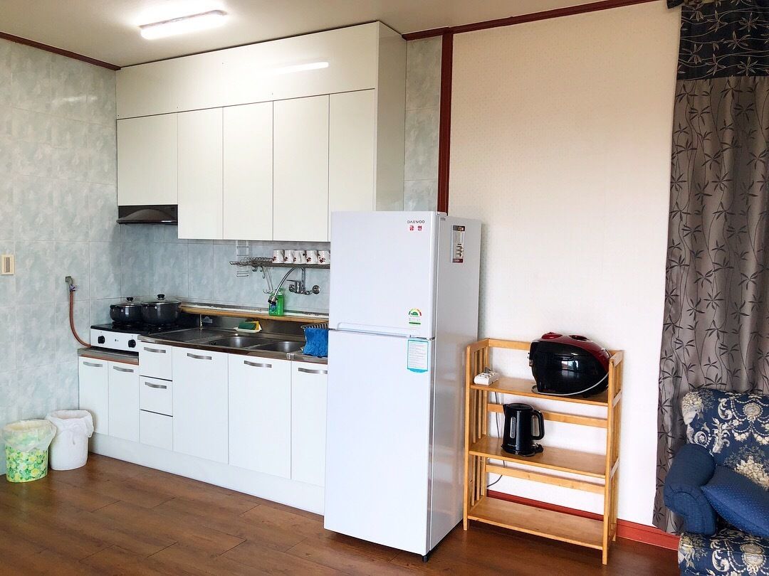 A compact kitchen with white cabinets, a built-in stove, and a refrigerator. A wooden shelf holds a coffee maker and a kettle.