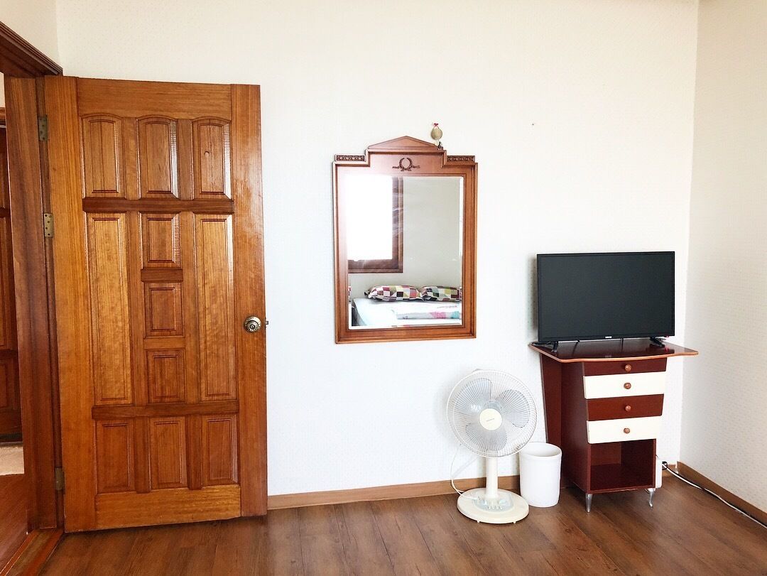 A room with a wooden door, a mirror, a television, a fan, and a dresser.