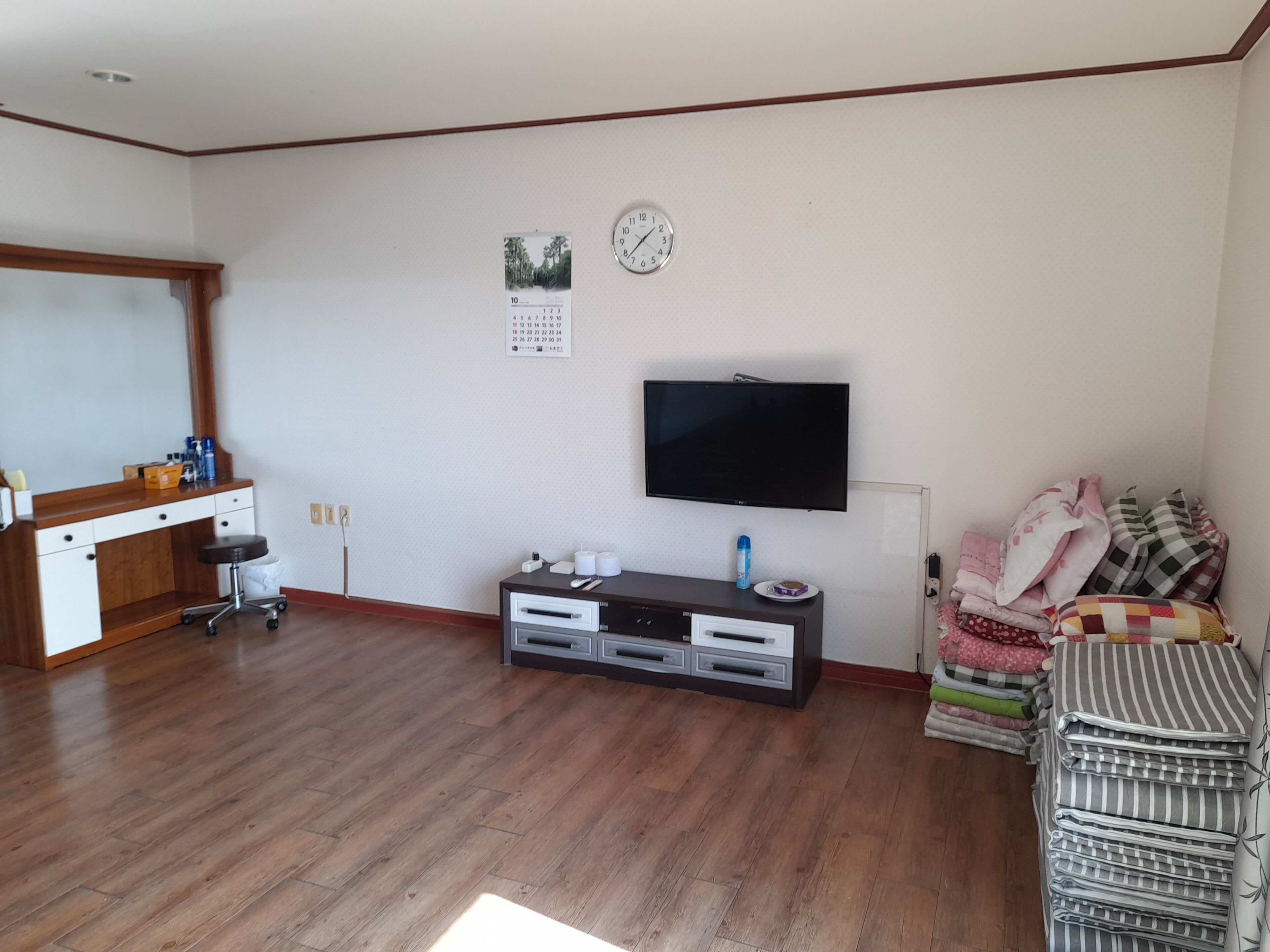 A living room with a wooden floor, a TV mounted on the wall, a desk with a chair, and a sofa with cushions.