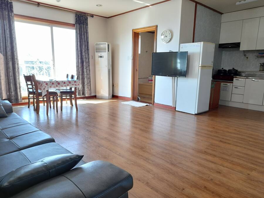Una sala de estar con sofá, mesa de comedor, sillas, aire acondicionado, televisor y un área de cocina con armarios y refrigerador.