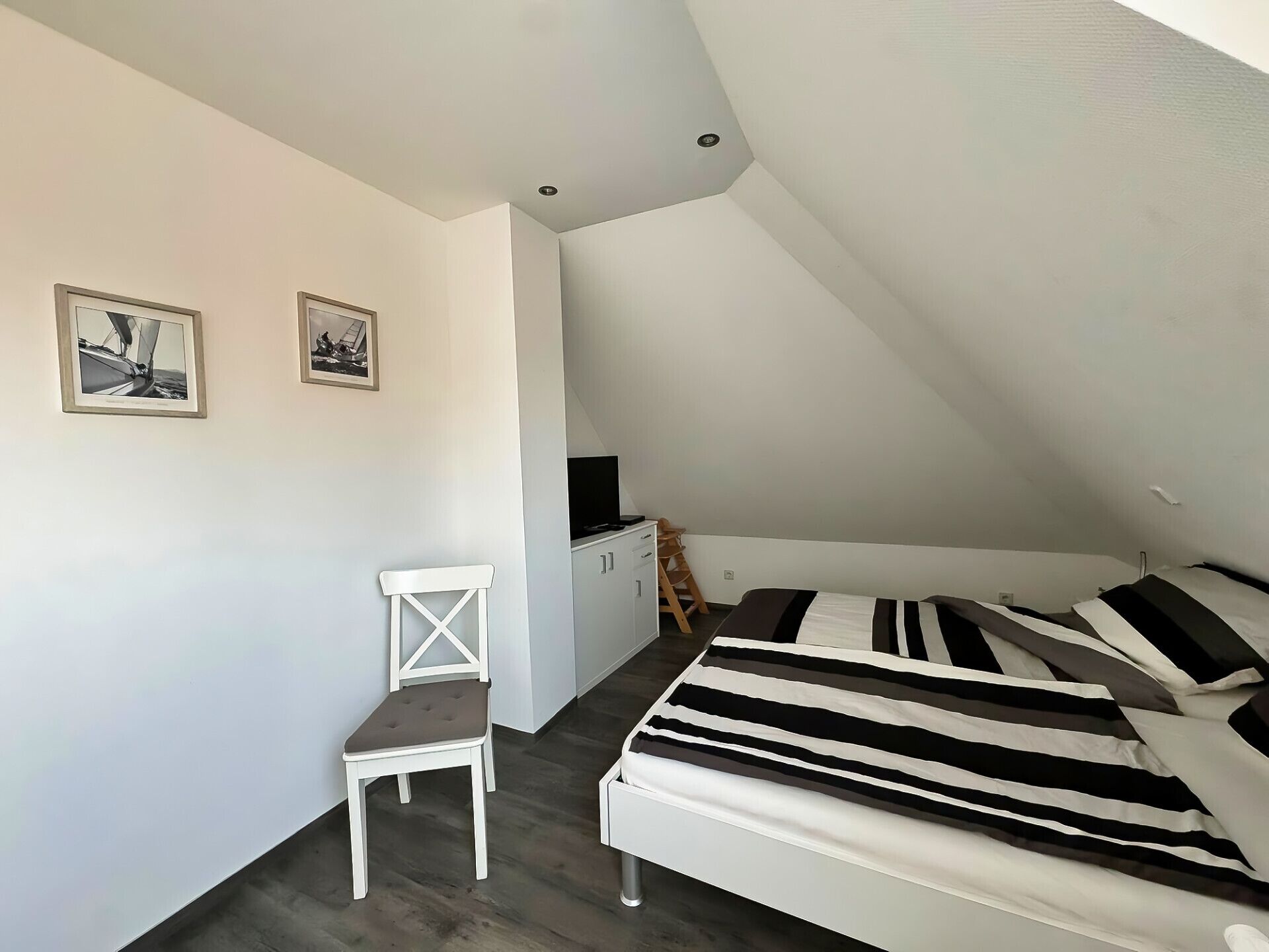 Un dormitorio pequeño y moderno con techo inclinado, cama con ropa de cama a rayas, una silla blanca y dos cuadros en la pared.