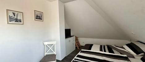 Un dormitorio pequeño y moderno con techo inclinado, cama con ropa de cama a rayas, una silla blanca y dos cuadros en la pared.