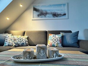 An attic room with a sofa, nautical-themed pillows, a painting of a beach, and jars with anchors on a tray.