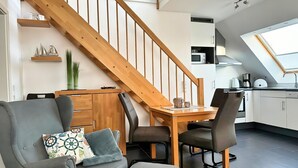 Un espacio habitable moderno con una escalera de madera, un área de comedor con mesa y sillas, una cocina con tragaluz y un sofá gris con cojines decorativos.