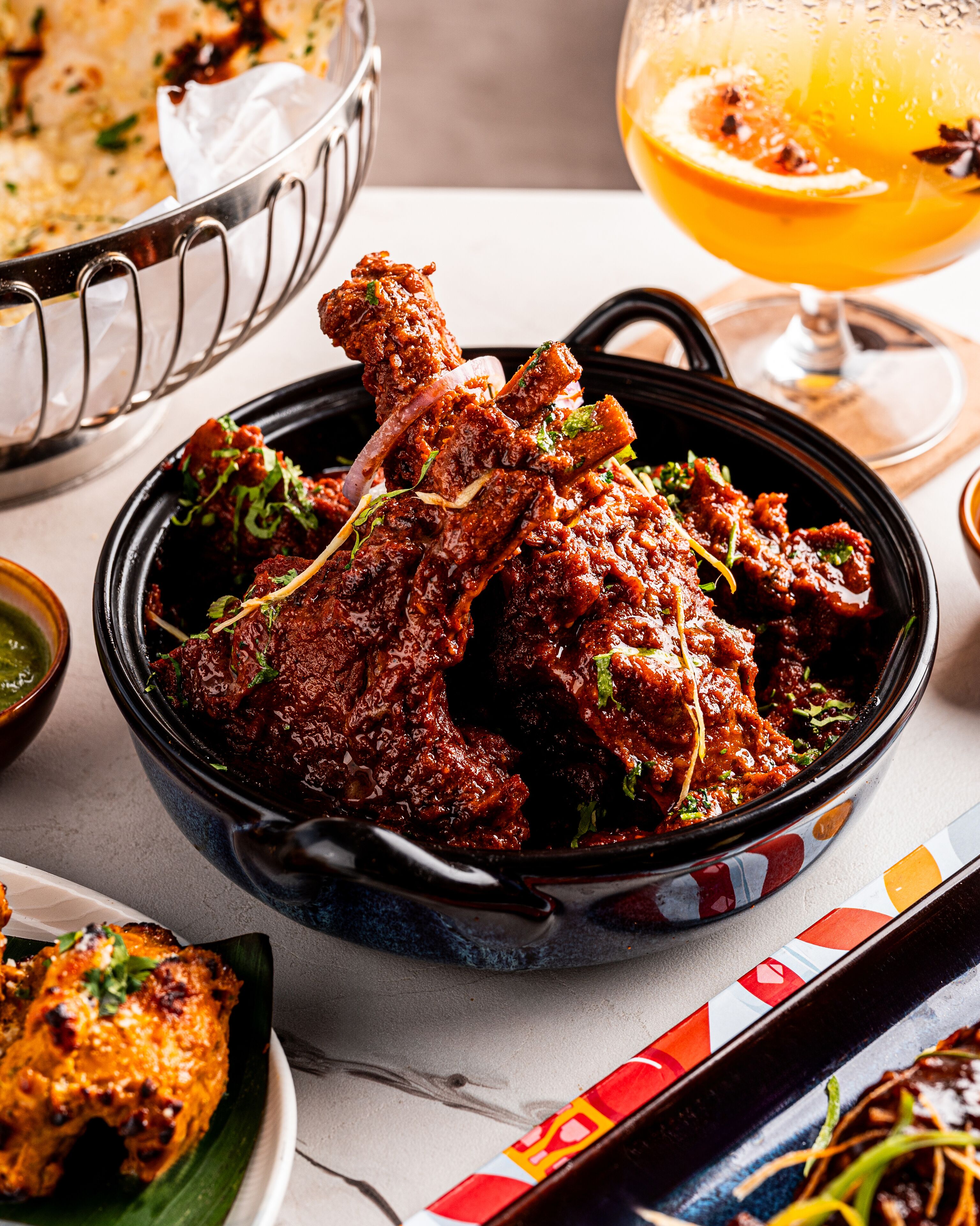 A bowl of spicy chicken wings garnished with herbs, a glass of orange juice, and a plate of fried food.