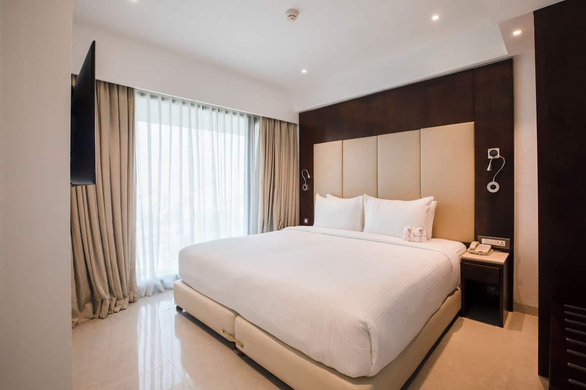 A hotel room with a large bed, a flat-screen TV mounted on the wall, a bedside table with a phone, and a window with curtains.