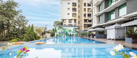 Una piscina con borde de azulejos, rodeada de vegetación y un edificio con balcones.