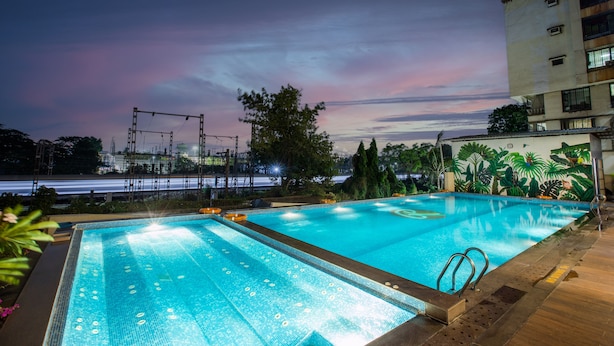 A rectangular outdoor swimming pool with a tiled edge, surrounded by greenery and a building. - Ramee Techome (Mumbai)
