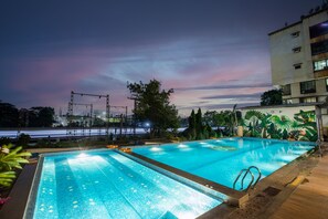 A rectangular outdoor swimming pool with a tiled edge, surrounded by greenery and a building.