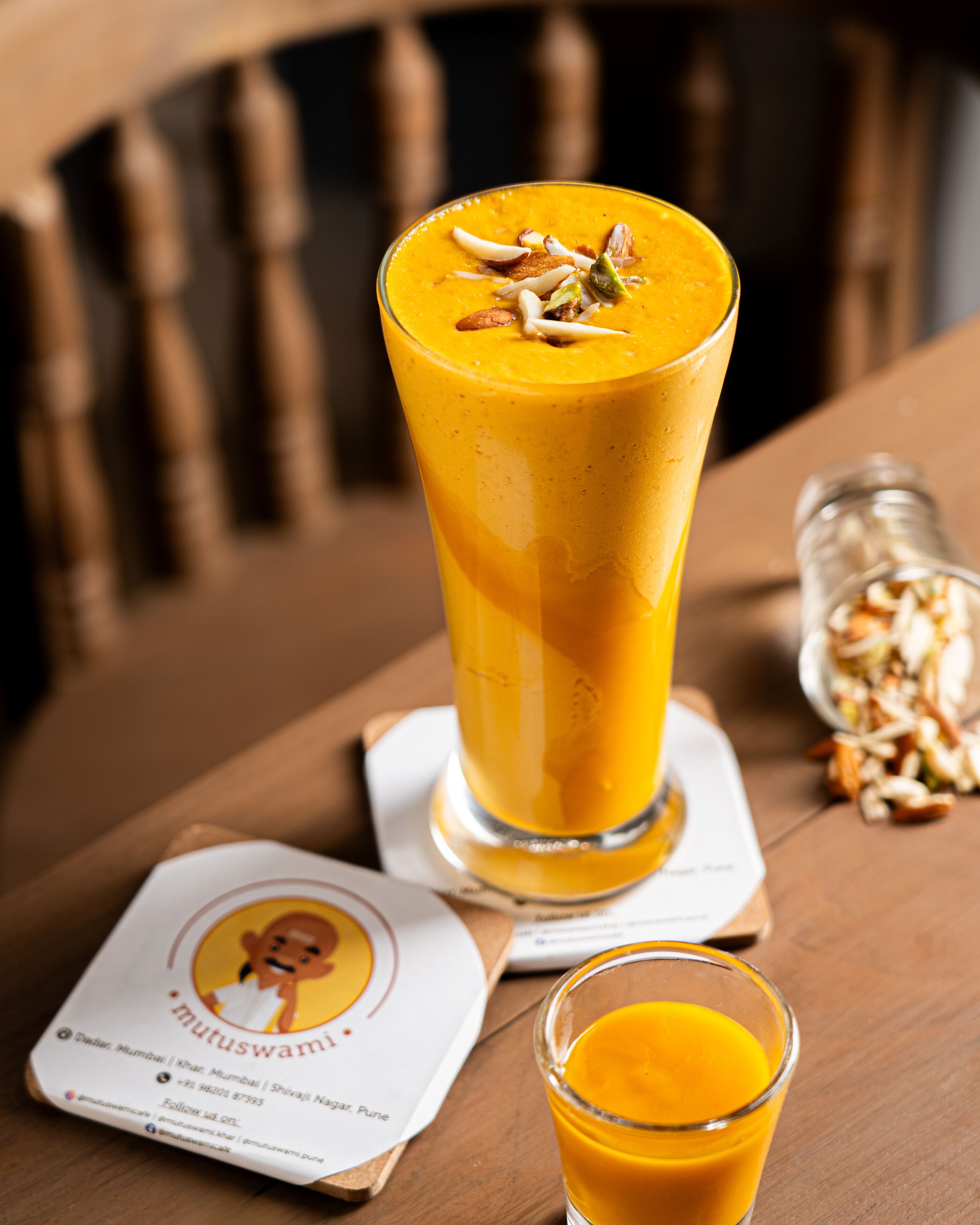 A tall glass of orange smoothie with nuts on top, placed on a wooden table with a coaster.