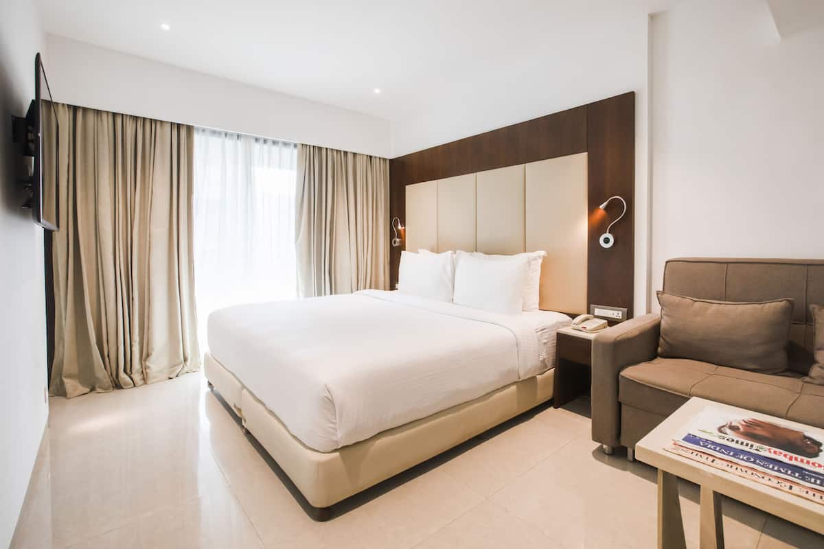 A hotel room with a large bed, a sofa, a TV, and a bedside table with a lamp.