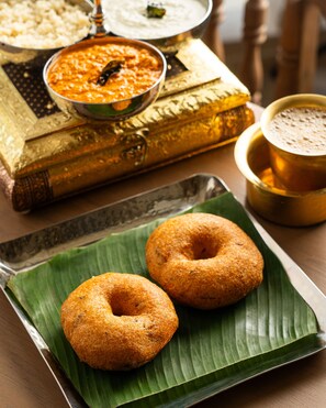 Two fried dough pastries on a banana leaf, a bowl of curry, and a cup of tea on a table with a book and decorative items in the background.