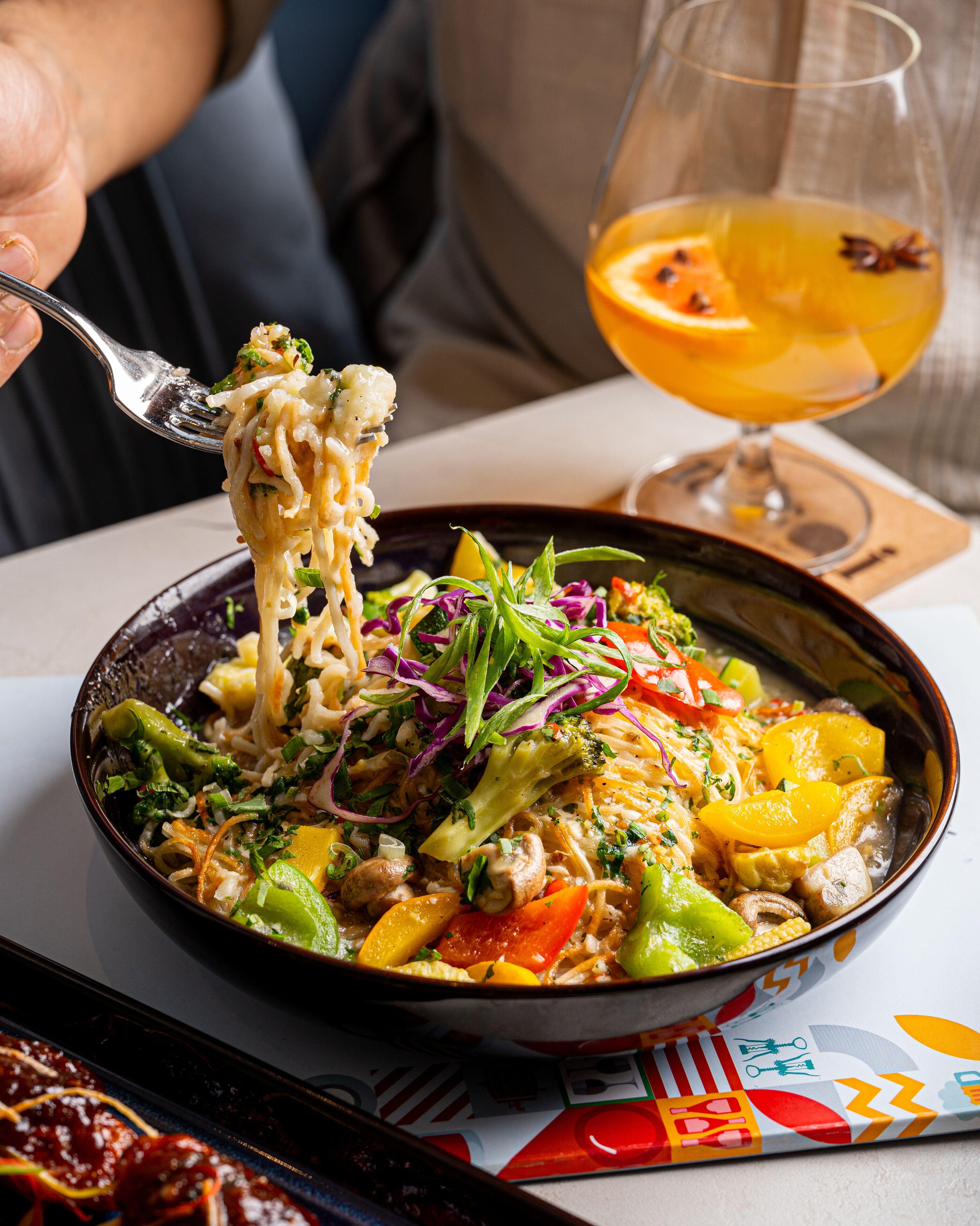 Un bol de pâtes aux légumes et un verre de jus d’orange sur une table.