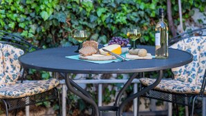 Une table ronde en métal, recouverte d’une nappe bleue, est entourée de chaises à motifs. Sur la table, on trouve une bouteille de vin blanc, un verre de vin blanc, un bol de raisins, un morceau de fromage, un couteau et une planche à découper en bois avec une tranche de pain.