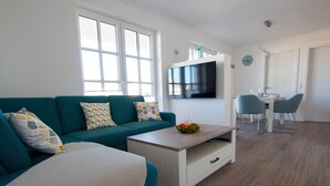 A modern living room with a teal sofa, a coffee table, and a TV mounted on the wall. - Apartment beach life. 2 bedrooms, high quality furnishings, great sea views! (Börgerende-Rethwisch)