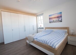 A bedroom with a bed, a nightstand, a window, and a wardrobe. - Apartment beach life. 2 bedrooms, high quality furnishings, great sea views! (Börgerende-Rethwisch)