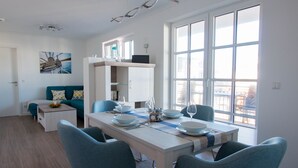 A modern, well-lit dining area with a white table set for four, blue chairs, and a view of the city through large windows. - Apartment beach life. 2 bedrooms, high quality furnishings, great sea views! (Börgerende-Rethwisch)