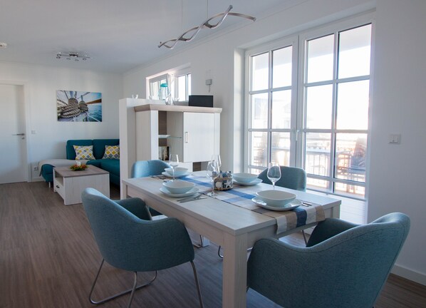 A modern, well-lit dining area with a white table set for four, blue chairs, and a view of the city through large windows. - Apartment beach life. 2 bedrooms, high quality furnishings, great sea views! (Börgerende-Rethwisch)