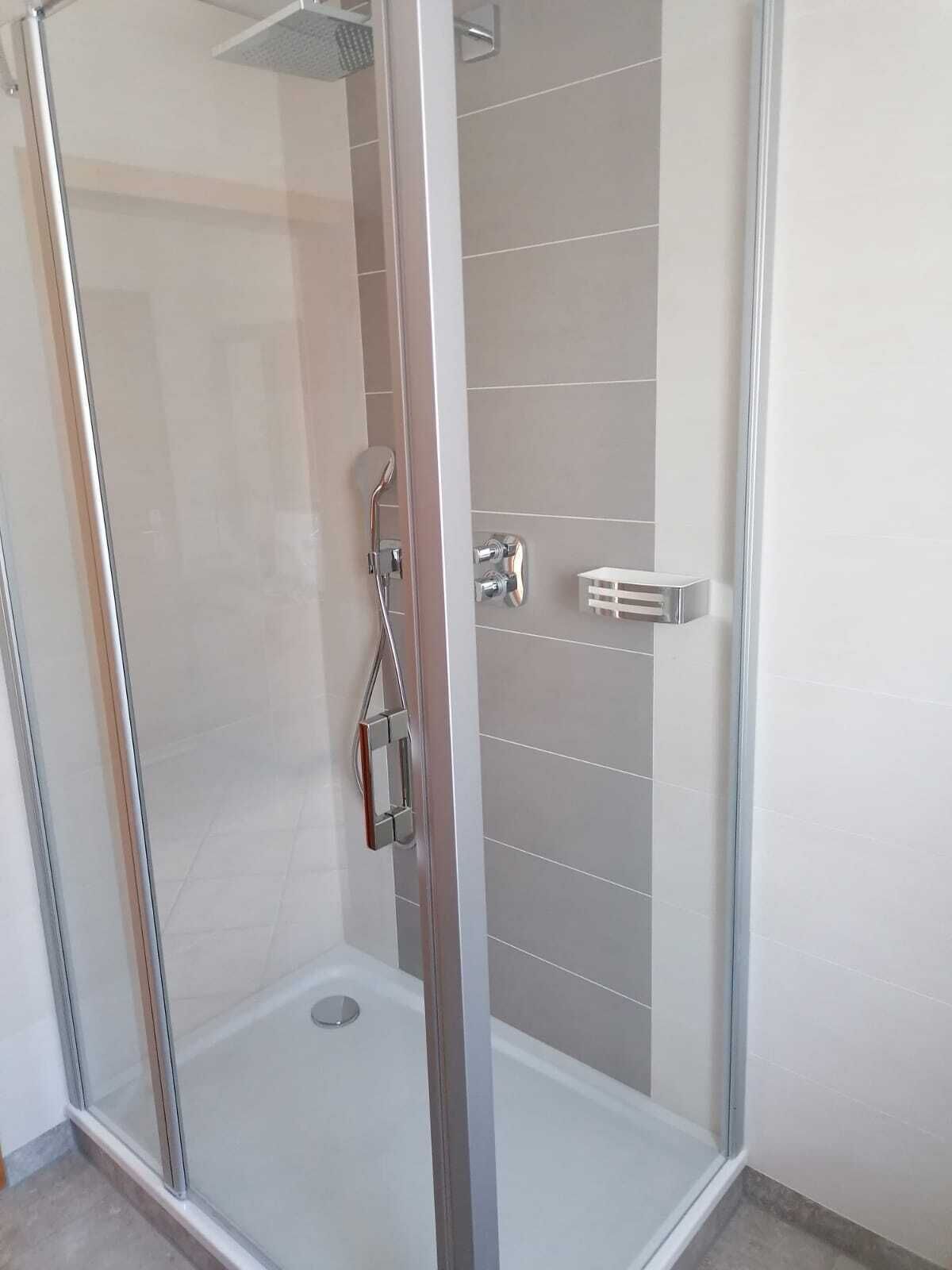 Une douche fermée par des parois en verre, dotée d’un pommeau de douche et d’un panneau de commande en chrome, d’un mur recouvert de carreaux blancs et d’une petite étagère blanche.