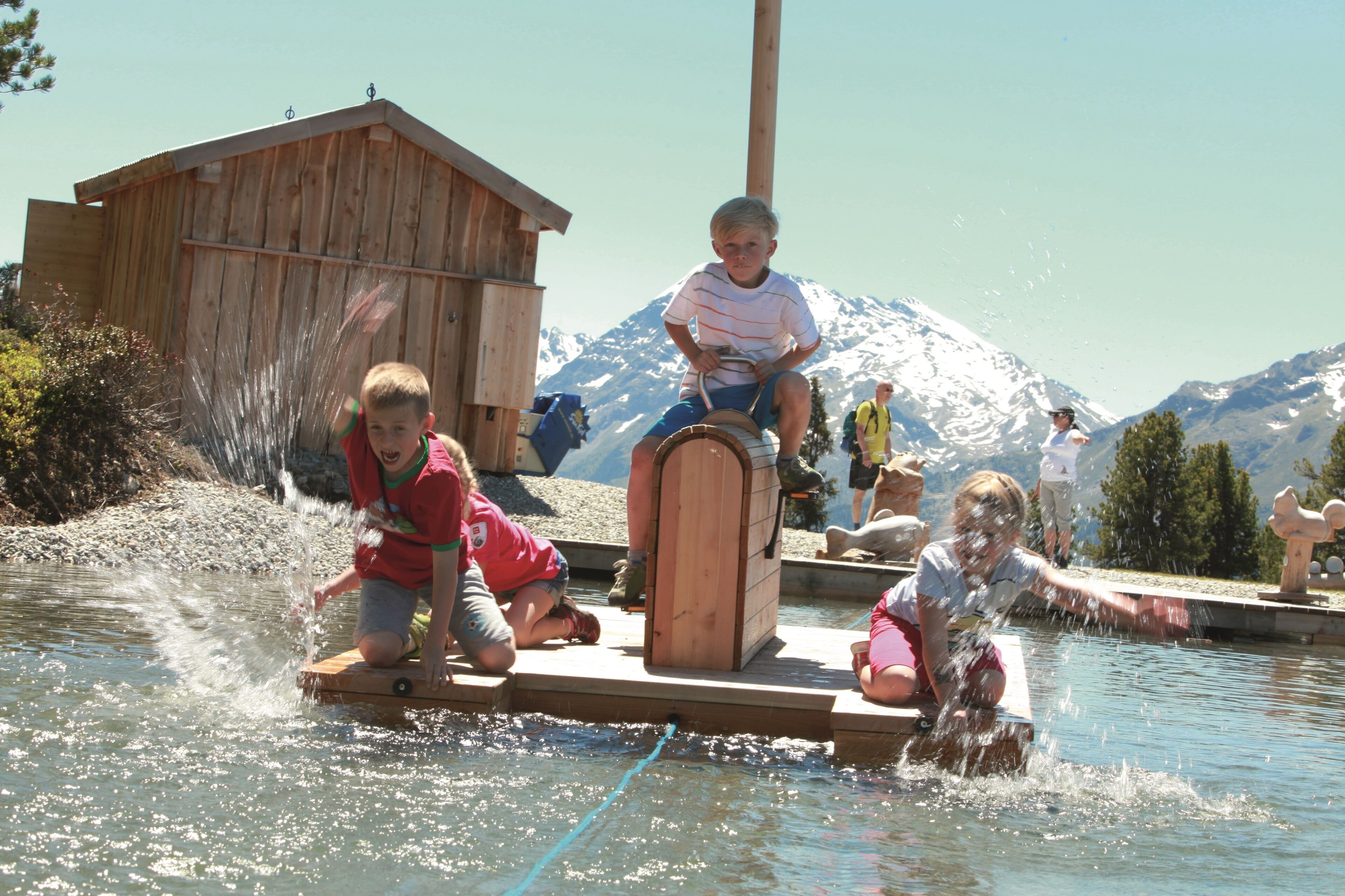 A wooden dock with people playing in the water, a wooden structure in the background, and snow-capped mountains.
