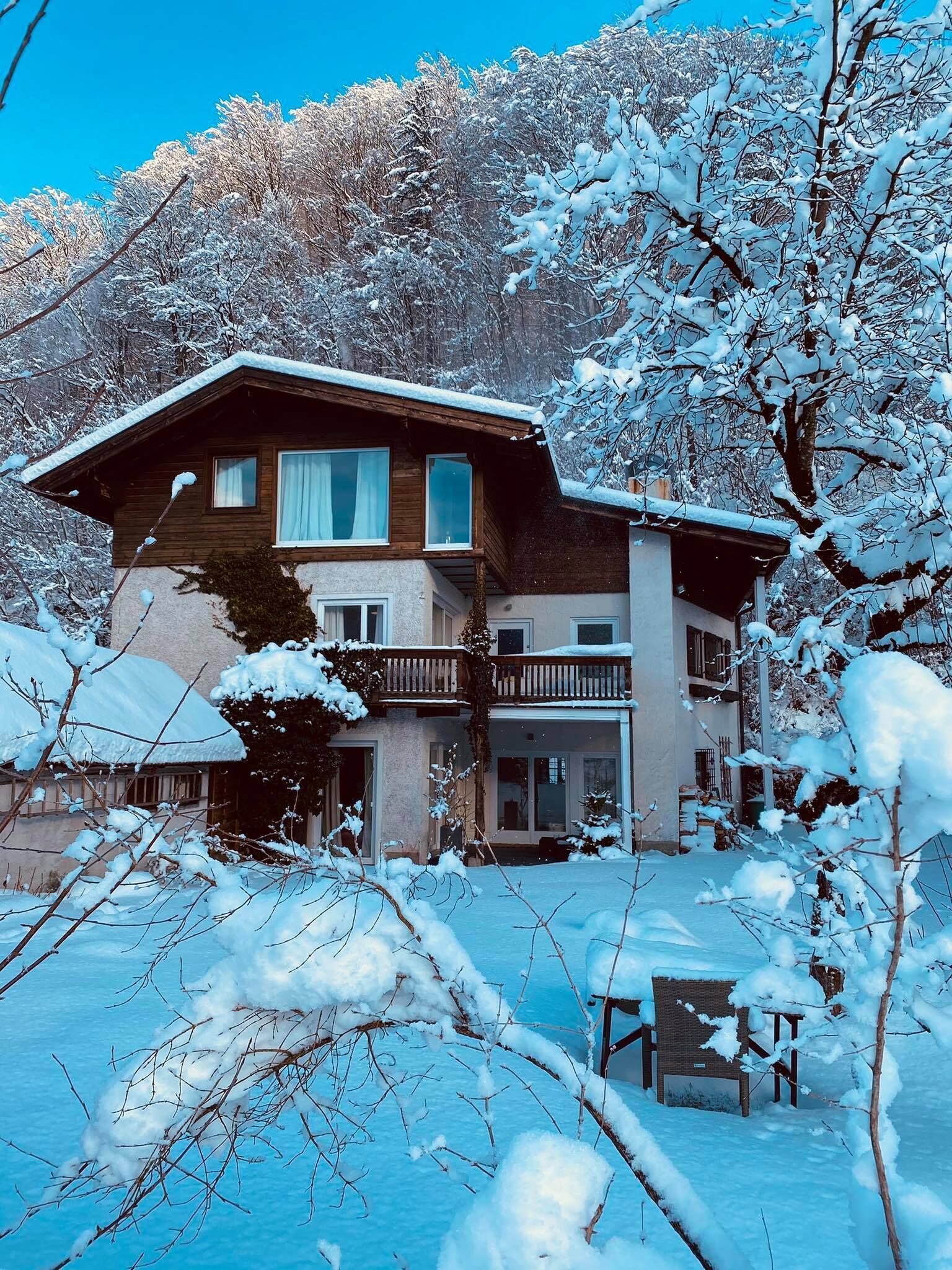 A two-story wooden house with a balcony, surrounded by snow-covered trees.