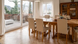 A dining area with a wooden table and chairs, a cabinet with glass doors, and a view through large windows.