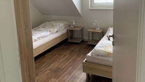 En liten lägenhet med ett enda rum, ett sluttande tak, trägolv, två sängar, ett fönster med persienner och ett litet nattduksbord med en lampa.