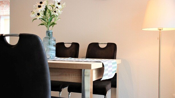 En matplats med ett bord, stolar, en vas med blommor, en klocka och en lampa.