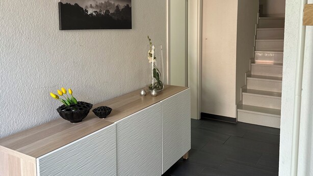 A modern interior with a wooden console table, a framed landscape painting, and a staircase leading to an upper level.