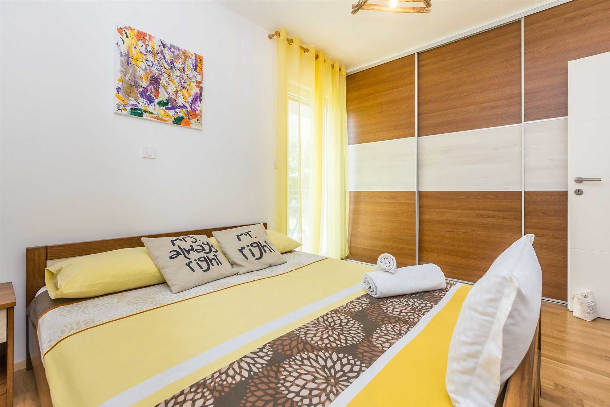 Un dormitorio con cama, almohadas, un armario de madera, una ventana con cortinas y un cuadro en la pared.