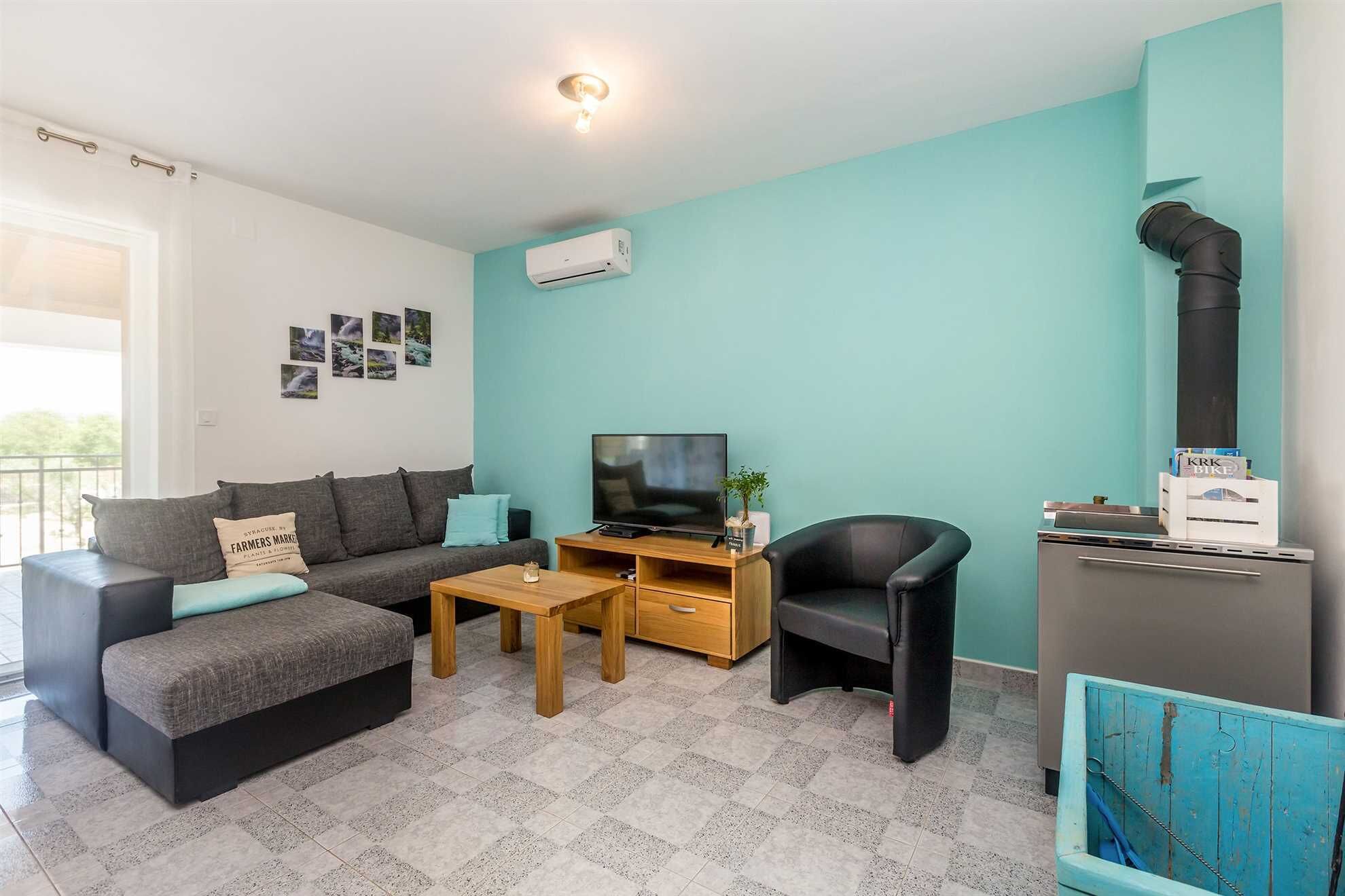 Uma sala de estar moderna com um sofá cinzento, uma mesa de centro de madeira, uma poltrona preta e uma TV. Há uma pequena planta sobre a mesa de centro e um porta-retrato na parede.