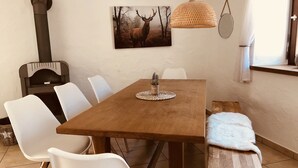 Un comedor con una mesa de madera, sillas blancas, una lámpara colgante de fibra y una chimenea, además de un cuadro de un ciervo en la pared.