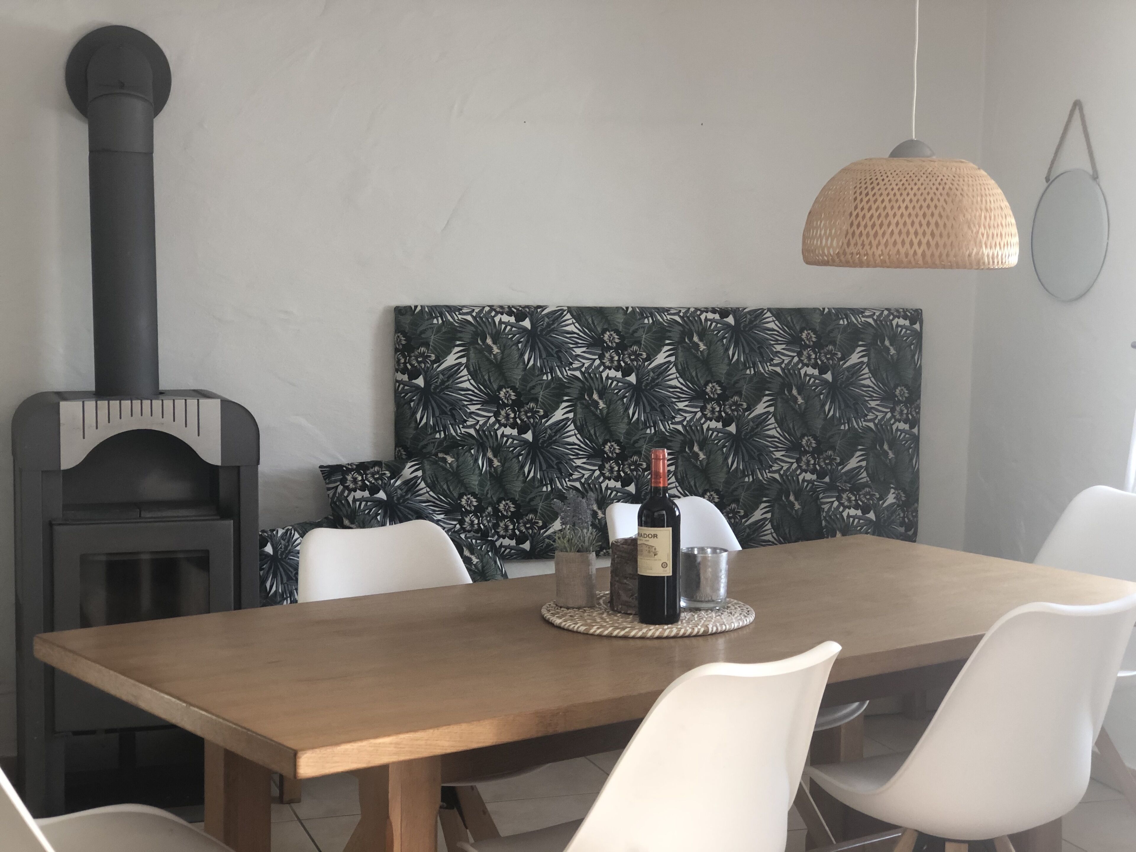 A dining area with a wooden table, white chairs, a woven pendant light, and a black wood-burning stove.