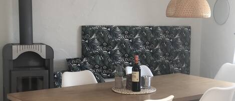 Un comedor con una mesa de madera, sillas blancas, una lámpara colgante tejida y un estufa de leña negra.