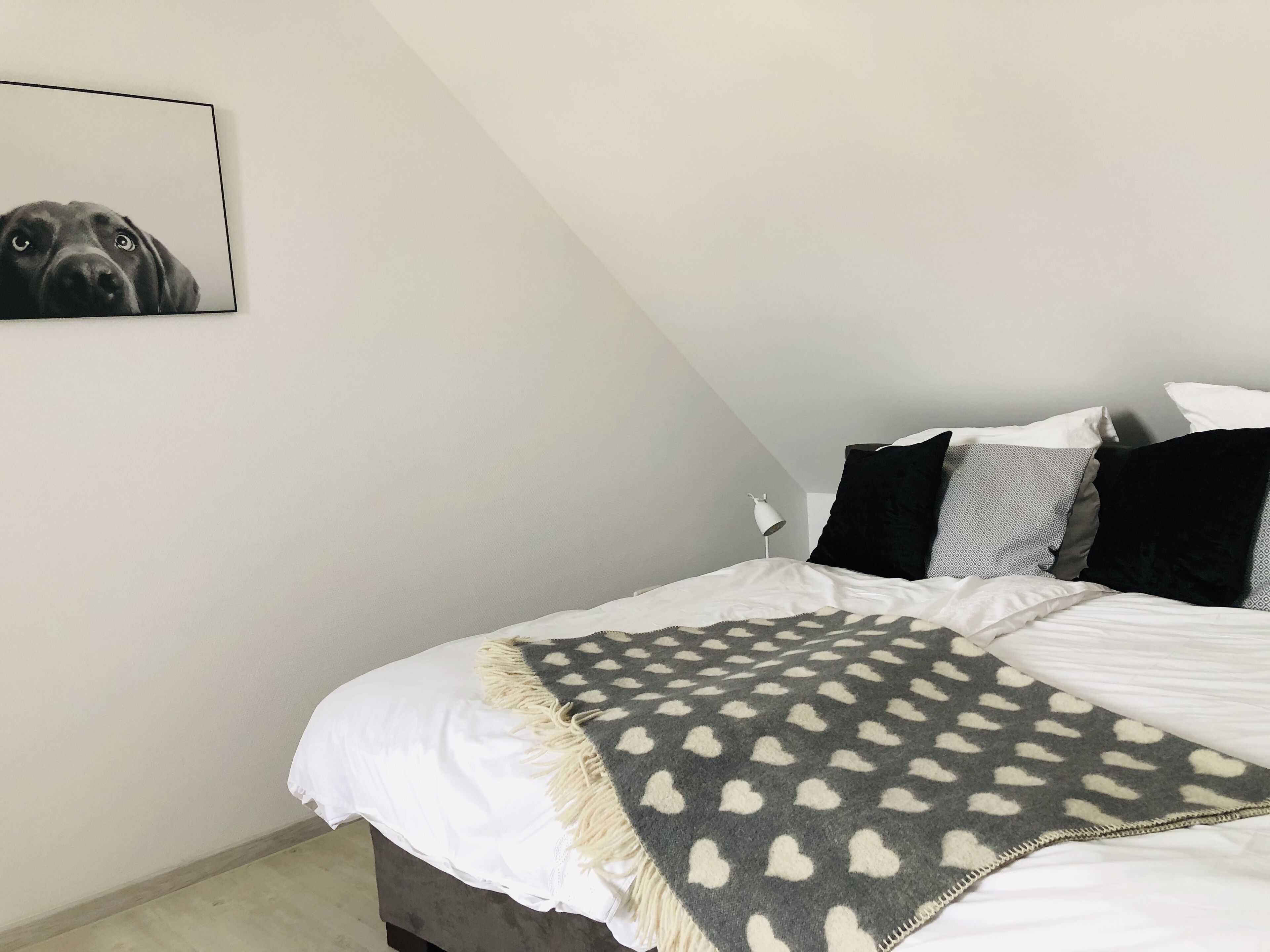 Une chambre à coucher avec un lit, des oreillers, un plaid, un tableau accroché au mur et une lampe de chevet.