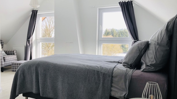 Un dormitorio moderno con una cama grande, una mesita redonda con una lámpara y dos ventanas con cortinas.