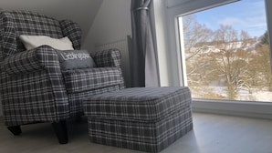 Una habitación con un sillón y reposapiés escoceses, un ventanal con vistas a árboles y edificios, y un radiador.