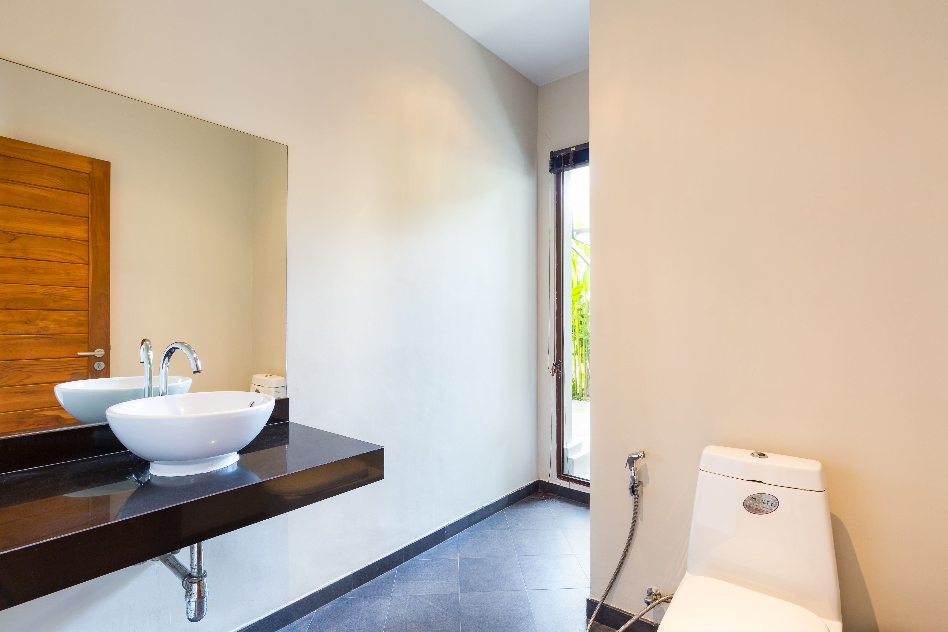 A modern bathroom with a black countertop, two white sinks, a wooden door, and a toilet.