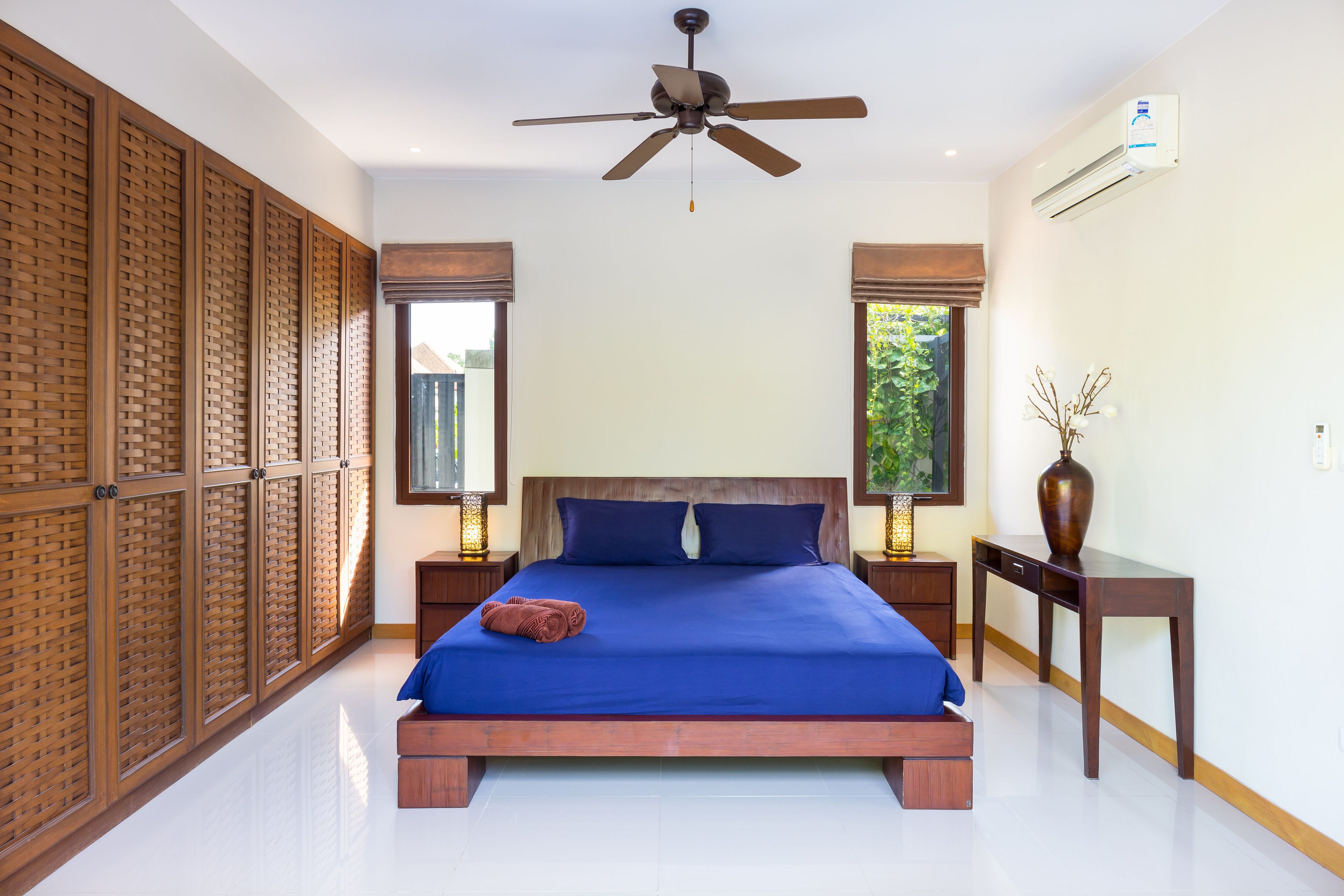 A bedroom with a large bed, wooden headboard, two bedside tables with lamps, a ceiling fan, and a window with a view of greenery.