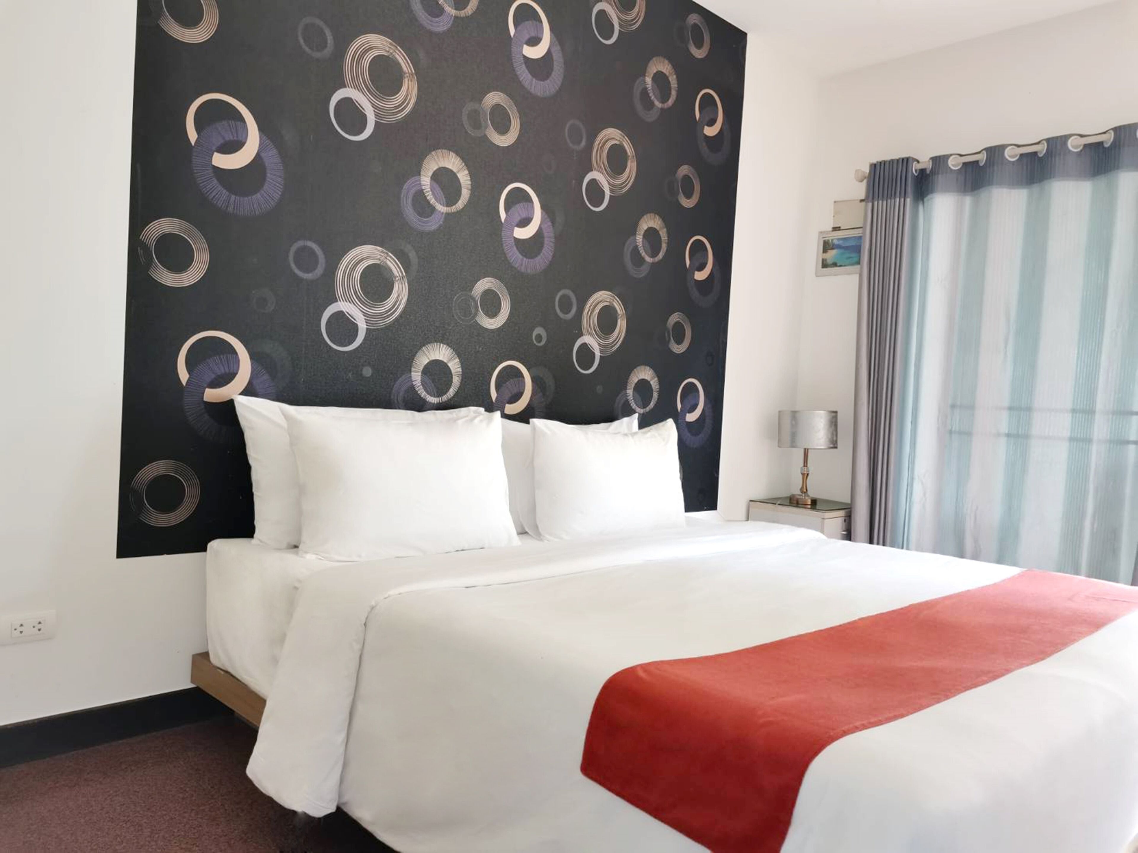 Una habitación de hotel con cama, mesita de noche, lámpara y ventana con cortinas.