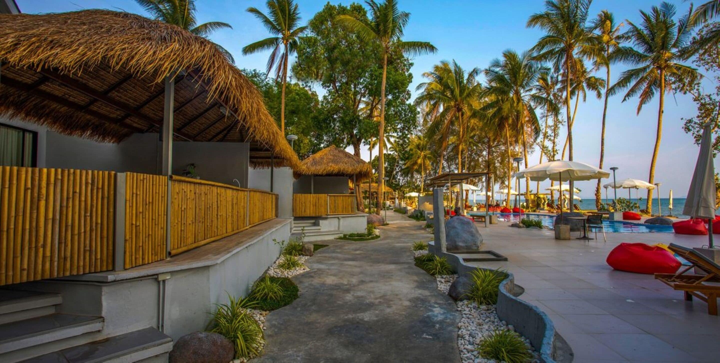 Un resort con construcciones de techo de paja, palmeras, un área de piscina con sillones de descanso y una vista despejada del océano.