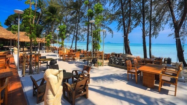 Un espace extérieur aménagé avec des tables et des chaises en bois, entouré d’arbres et offrant une vue sur la plage.