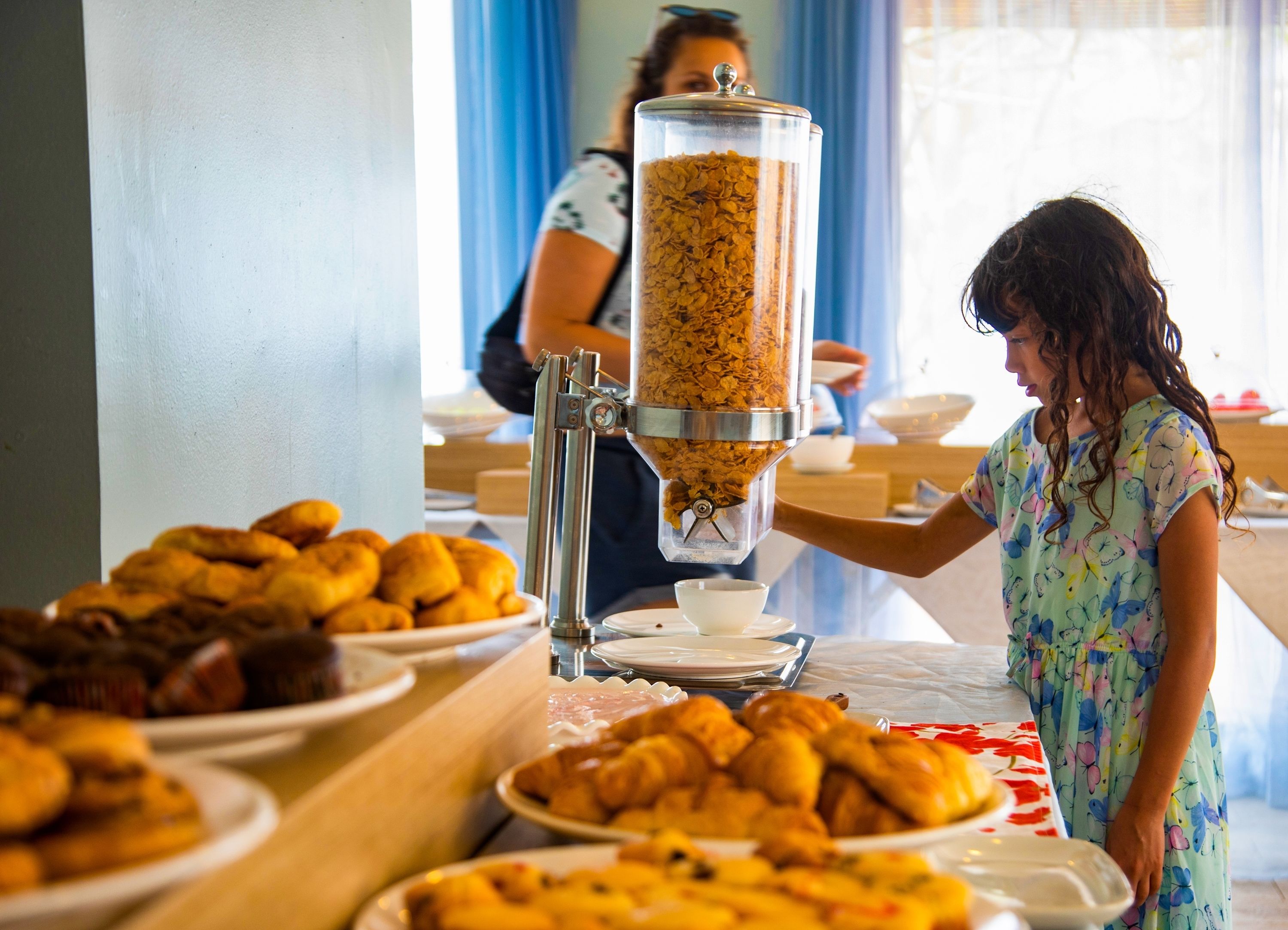 A buffet setup with a cereal dispenser, plates of pastries, and a person serving food.