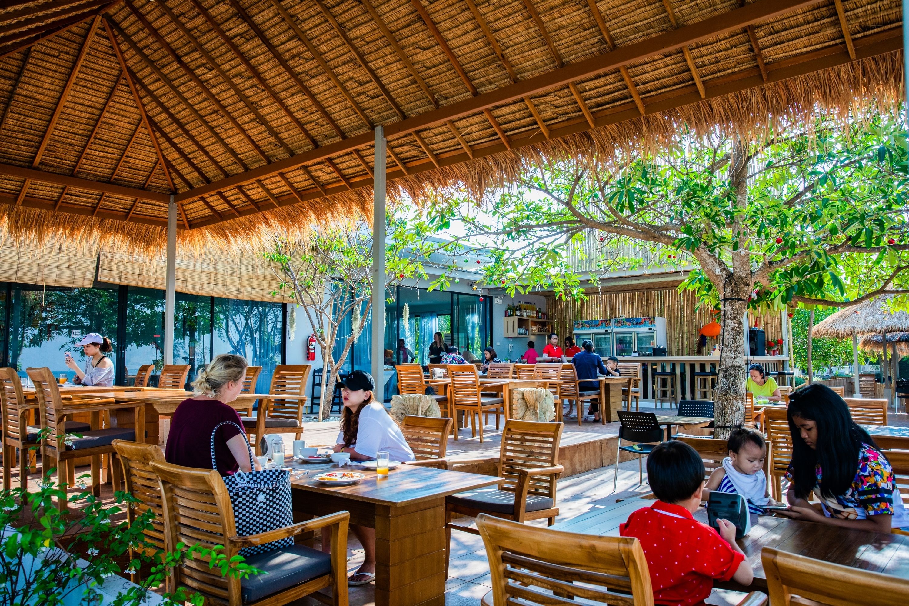 An outdoor dining area with wooden tables and chairs, a thatched roof, and a view of the surrounding greenery.